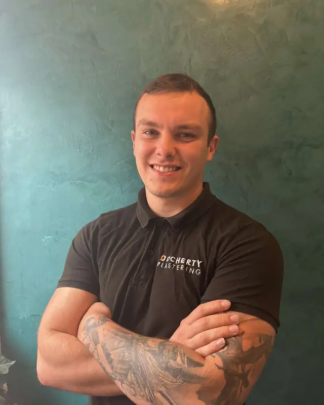 Liam Docherty, smiling with his arms folded, in a black Docherty Plastering polo shirt in front of a teal polished plaster wall.