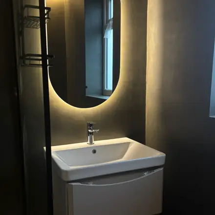 A wall-hung basin and backlit oval mirror set against a warm grey seamless plastered wall, with no joints or tile lines.